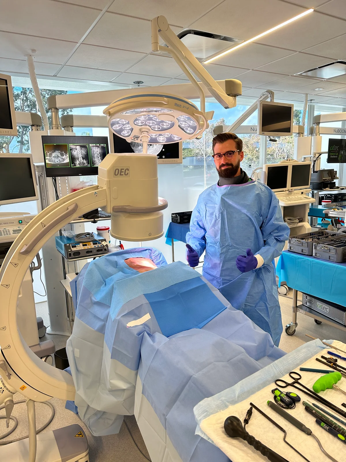 Tim wearing a blue gown and gloves beside a draped table, imaging equipment, and surgical instruments.