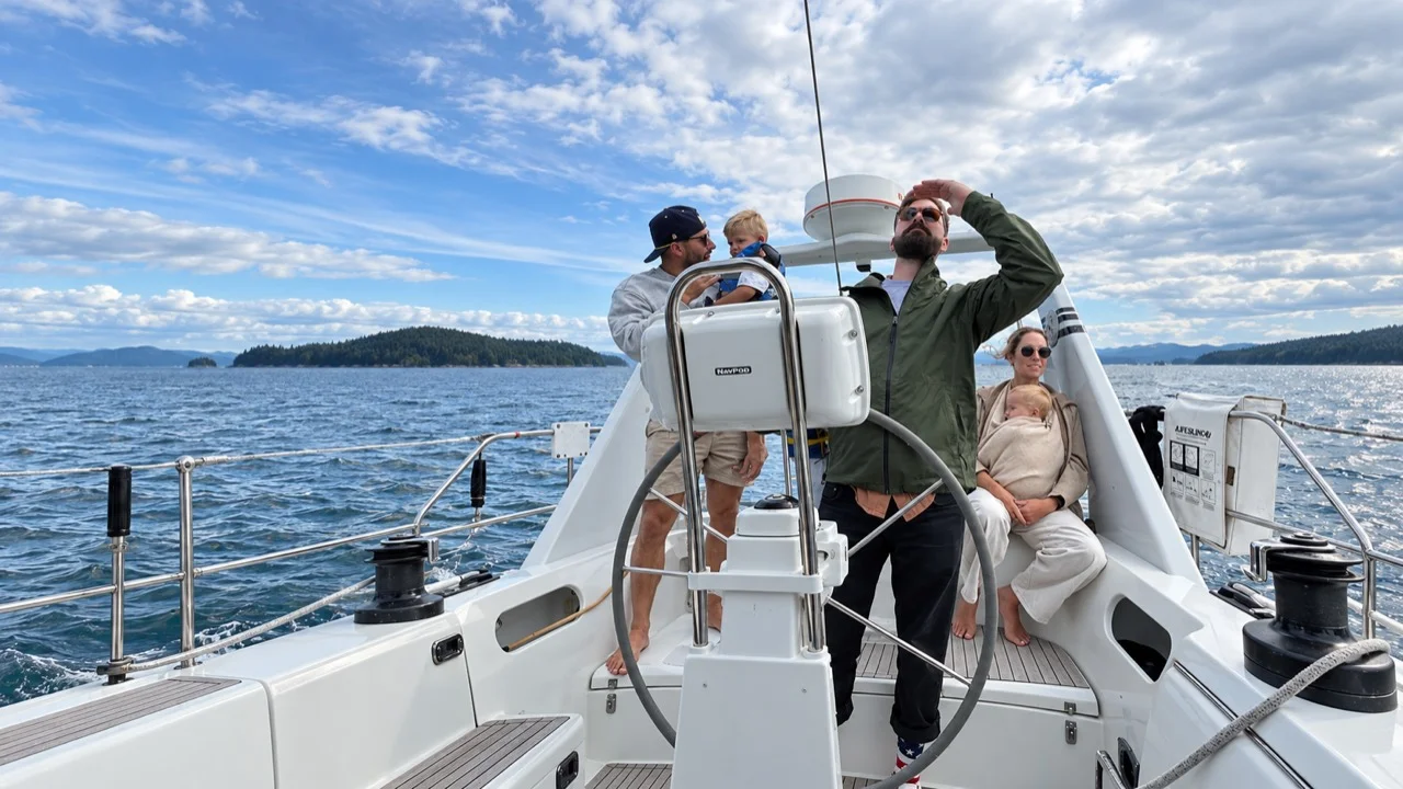 A group of adults and children aboard a sailboat on open water near wooded islands.